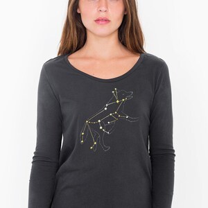 May include: Dark gray long-sleeve shirt with a scoop neck. The shirt features a constellation design of a dog, with white and yellow star accents. The dog constellation is on the front of the shirt.