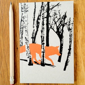 May include: A small, brown notebook featuring a black and white illustration of a fox walking through a forest. The fox is orange and the trees are silhouetted against a white background.