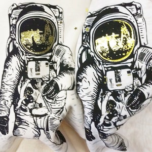 Peut inclure: Deux illustrations en noir et blanc d'astronautes portant des casques avec des accents en feuille d'or. Les astronautes portent des combinaisons spatiales et se tiennent sur un fond blanc.