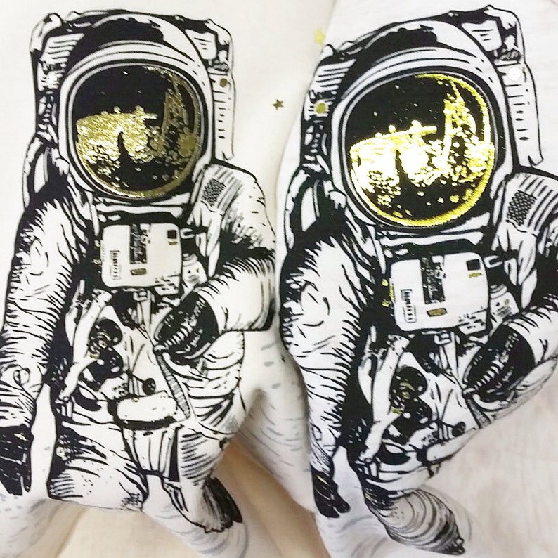 Peut inclure: Deux illustrations en noir et blanc d'astronautes portant des casques avec des accents en feuille d'or. Les astronautes portent des combinaisons spatiales et se tiennent sur un fond blanc.