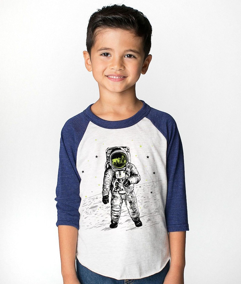 Peut inclure: Un t-shirt de baseball blanc et bleu avec un graphique d'un astronaute sur la lune. L'astronaute porte une combinaison spatiale et un casque. Le t-shirt a une manche bleu marine et une manche blanche.
