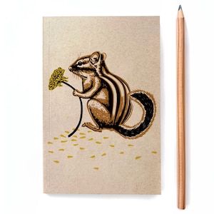 May include: A notebook with a chipmunk illustration holding a yellow flower. The chipmunk is brown and black with a striped pattern. A wooden pencil is next to the notebook on a white background.