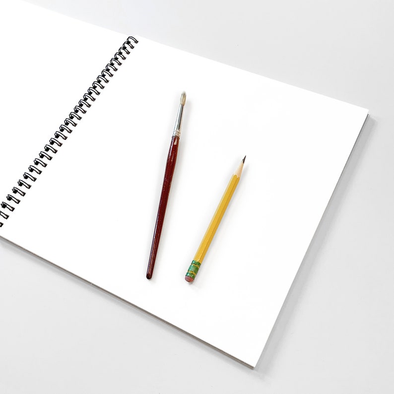 May include: A white spiral bound sketchbook with a yellow pencil and a brown paintbrush lying on top of it.