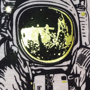 Peut inclure: Une illustration en noir et blanc d'un astronaute avec une lune en feuille d'or dans le casque. L'astronaute porte une combinaison spatiale avec une fermeture &eacute;clair et un patch sur le bras.