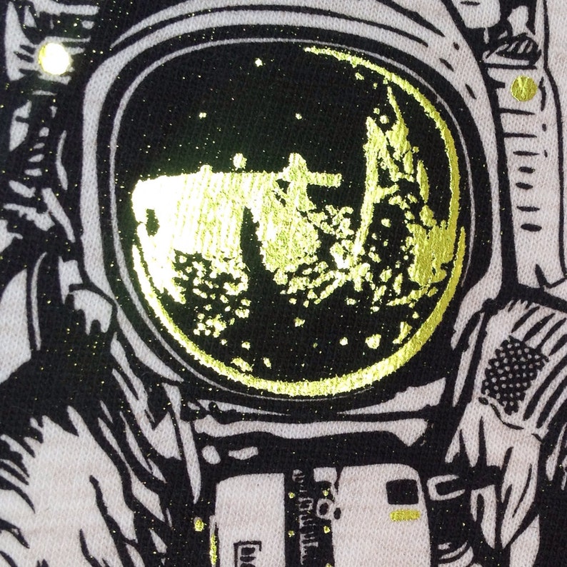 Peut inclure: Une illustration en noir et blanc d'un astronaute avec une lune en feuille d'or dans le casque. L'astronaute porte une combinaison spatiale avec une fermeture &eacute;clair et un patch sur le bras.