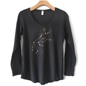 May include: Black long-sleeve shirt with a scoop neck. The shirt features a gold and white constellation design of a wolf. The wolf design is made of small stars connected by lines. The shirt is hanging on a wooden hanger.