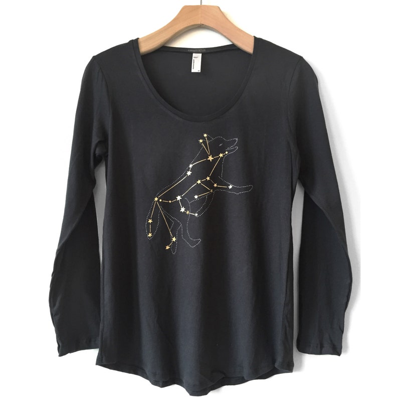 May include: Black long-sleeve shirt with a scoop neck. The shirt features a gold and white constellation design of a wolf. The wolf design is made of small stars connected by lines. The shirt is hanging on a wooden hanger.