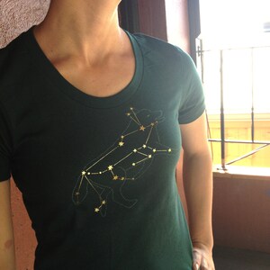 May include: A dark green t-shirt with a gold embroidered constellation design of a wolf. The design is detailed and intricate, with each star in the constellation clearly visible.
