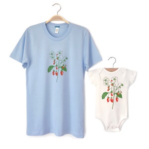 May include: A light blue t-shirt and a white bodysuit with a graphic print of a strawberry plant with red strawberries and white flowers.
