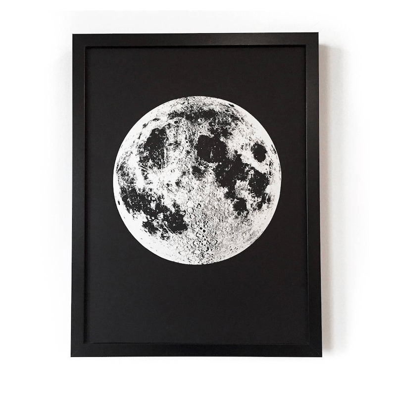 May include: A black framed print of a white moon on a black background. The moon is a detailed illustration of the lunar surface.
