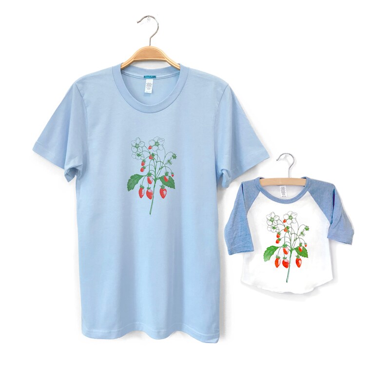 May include: Two light blue t-shirts with a white strawberry graphic. The larger shirt has short sleeves and the smaller shirt has a blue and white baseball style sleeve.
