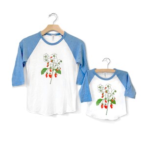 May include: Two baseball style t-shirts with a white base and blue sleeves. The shirts feature a graphic of a strawberry plant with red strawberries and white flowers.