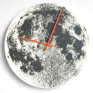 May include: A round wall clock with a white background and a black and white image of the moon. The clock has orange hands.