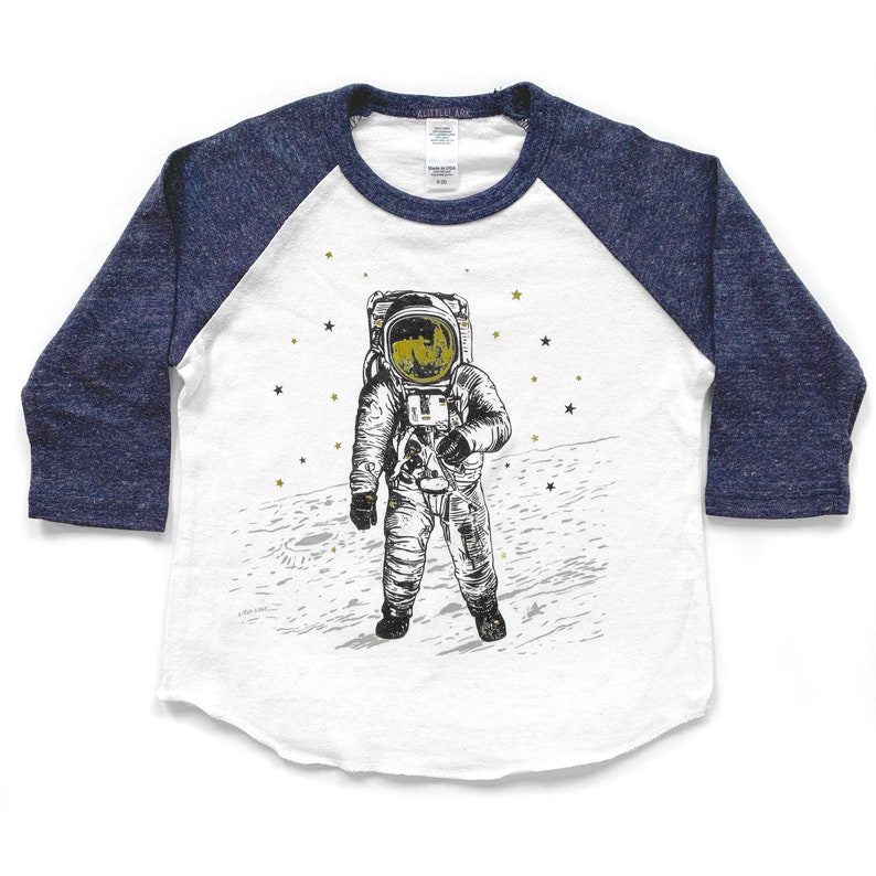 Peut inclure: Un t-shirt de baseball blanc et bleu avec un graphique d'un astronaute debout sur la lune. L'astronaute porte une combinaison spatiale blanche avec un casque dor&eacute; et est entour&eacute; d'&eacute;toiles dor&eacute;es.