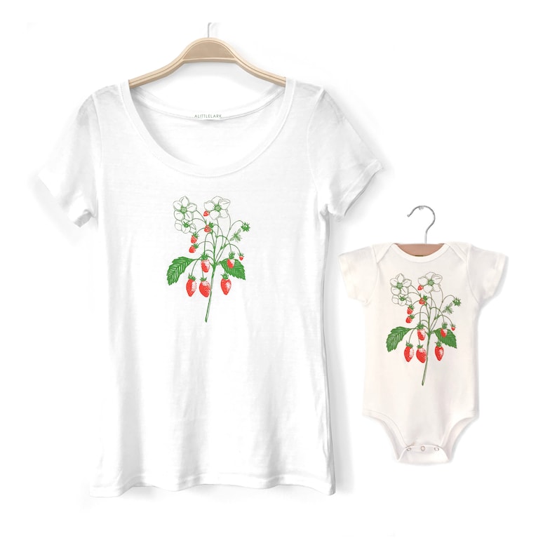 May include: A white t-shirt with a green and red strawberry plant design and a white onesie with the same design. The t-shirt has the word "Little Lark" printed on the inside of the neck.
