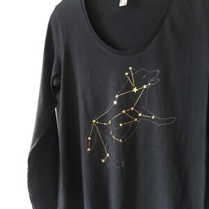 May include: A dark gray long-sleeve shirt with a scoop neck. The shirt features a gold constellation design of a wolf. The wolf is outlined with dotted lines and connected by gold lines and stars.