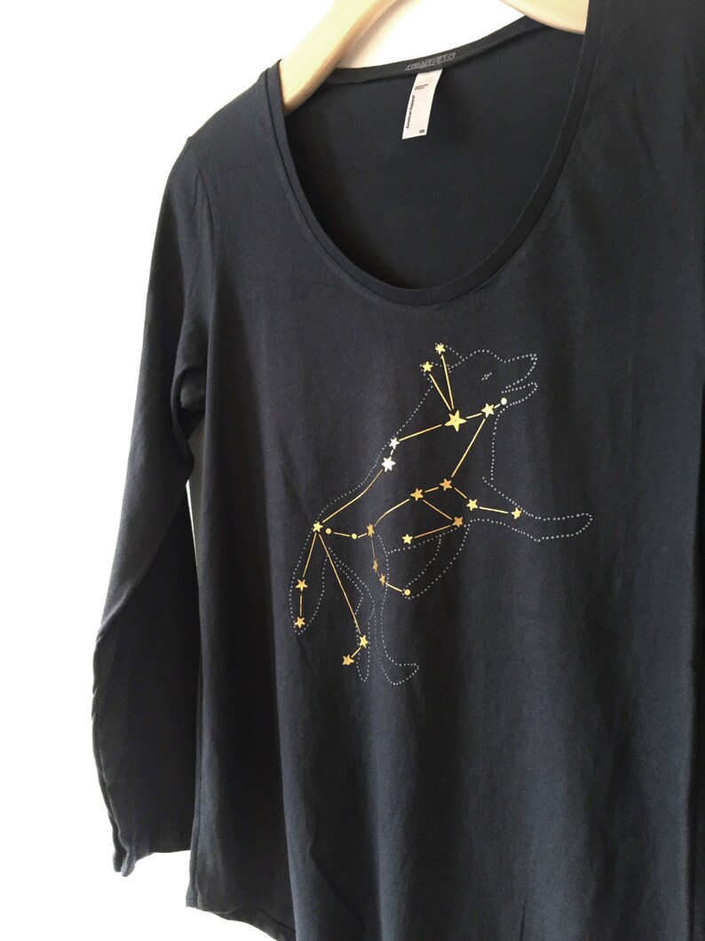 May include: A dark gray long-sleeve shirt with a scoop neck. The shirt features a gold constellation design of a wolf. The wolf is outlined with dotted lines and connected by gold lines and stars.
