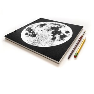 May include: A black spiral bound notebook with a white image of the moon on the front cover. Two pencils are lying on a white surface next to the notebook.
