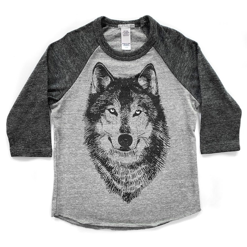 Puede incluir: Una camiseta de b&eacute;isbol gris y negra con un gr&aacute;fico de lobo blanco y negro en la parte delantera. La camiseta tiene un dise&ntilde;o de manga raglan.