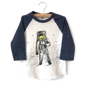 Peut inclure: Un t-shirt &agrave; manches longues blanc et bleu marine avec un motif d'astronaute sur la lune. L'astronaute porte une combinaison spatiale blanche avec un casque jaune et marche sur la surface de la lune. Le t-shirt a un design &agrave; manches raglan.