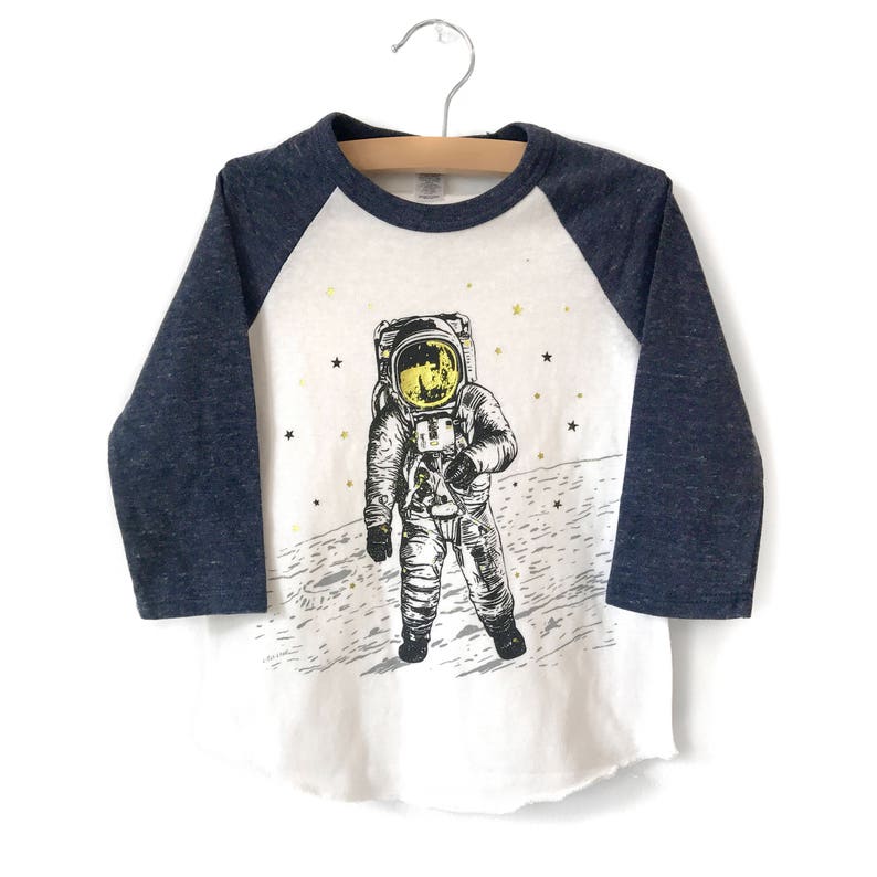 Peut inclure: Un t-shirt &agrave; manches longues blanc et bleu marine avec un motif d'astronaute sur la lune. L'astronaute porte une combinaison spatiale blanche avec un casque jaune et marche sur la surface de la lune. Le t-shirt a un design &agrave; manches raglan.