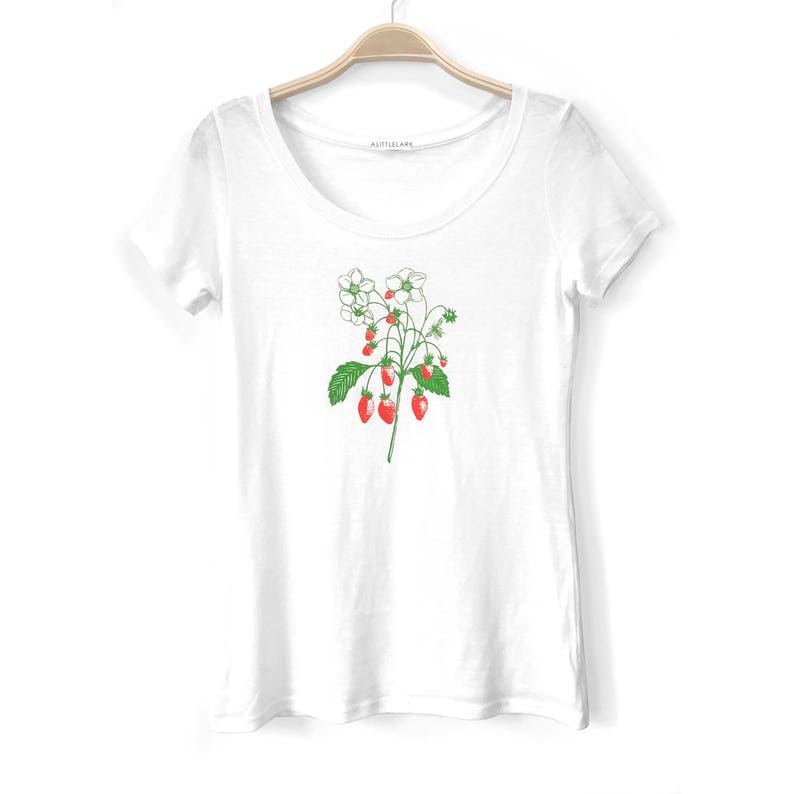 May include: White t-shirt with a green and red illustration of a strawberry plant with white flowers and red strawberries.