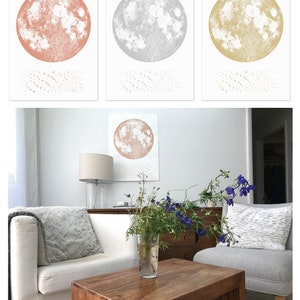 May include: Three framed prints of the moon in different colors: rose gold, silver, and gold. The prints are hanging on a white wall in a living room setting. A white couch, a wooden coffee table, and a vase of flowers are in the foreground.