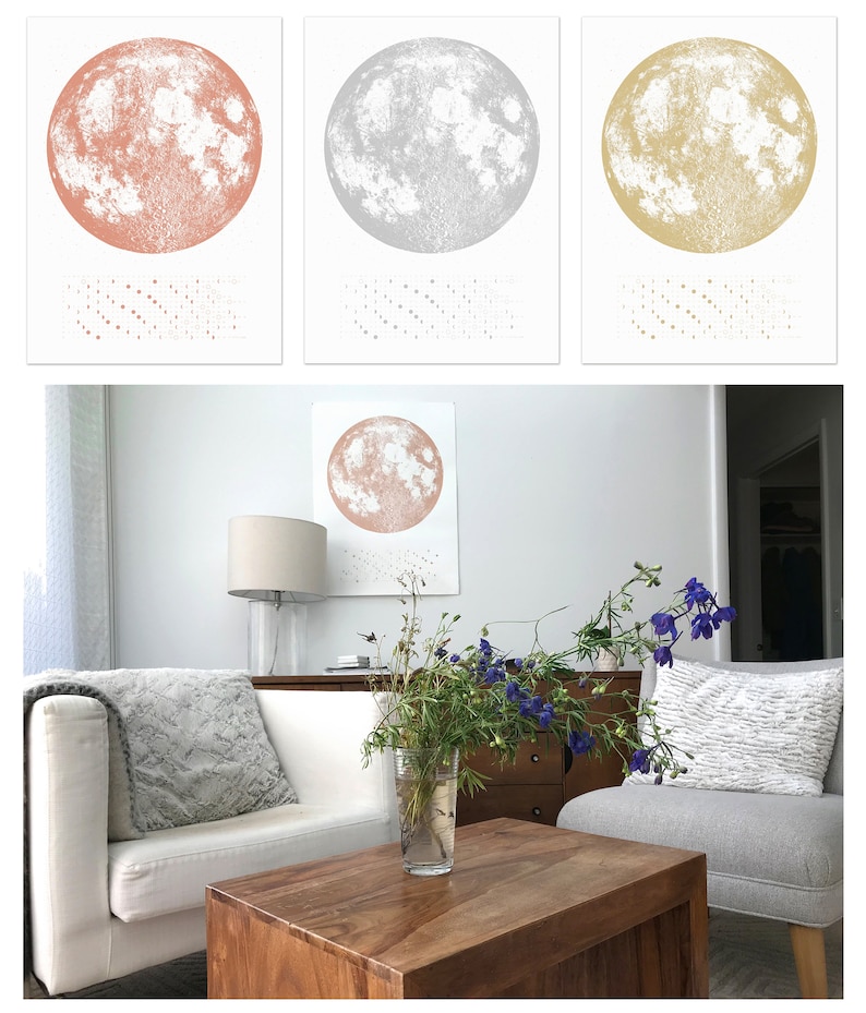 May include: Three framed prints of the moon in different colors: rose gold, silver, and gold. The prints are hanging on a white wall in a living room setting. A white couch, a wooden coffee table, and a vase of flowers are in the foreground.