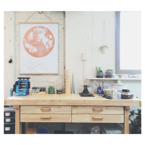 May include: A light wood workbench with three drawers, a blue metal rolling mill, and various tools and supplies. A poster with a large orange moon print hangs on the wall behind the workbench.