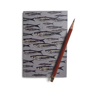 Peut inclure: Un carnet gris avec un motif d'illustrations de poissons noirs. Un crayon marron avec une virole et une gomme dorées repose sur le carnet. Le carnet et le crayon sont sur fond blanc.