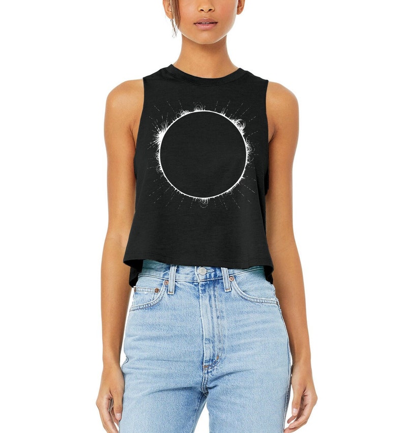 Woman is wearing a black, racerback cropped tank top printed with a total solar eclipse design on the front in white ink. Shirt by Little Lark