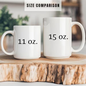 May include: Two white ceramic mugs, one labeled "11 oz." and the other "15 oz.", are displayed side-by-side for size comparison. The mugs have a standard handle design and are set on a wooden surface. The text "SIZE COMPARISON" is at the top.