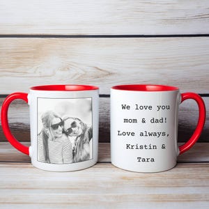 May include: Two white ceramic mugs with red interiors and handles. One mug features a black and white photo of two people wearing sunglasses. The other mug has the text "We love you mom & dad! Love always, Kristin & Tara."