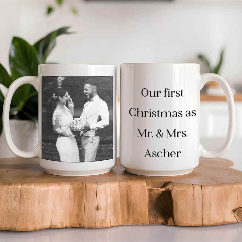 May include: Two white ceramic mugs. One mug features a black and white photo of a couple, the other mug has the text "Our first Christmas as Mr. & Mrs. Ascher" in black. Both mugs have large handles.
