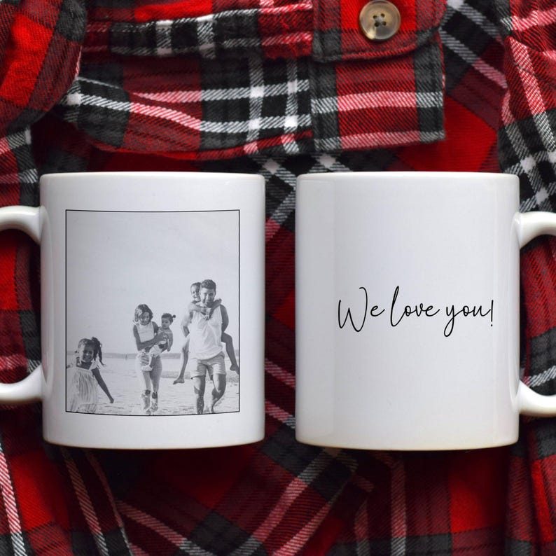 May include: Two white ceramic mugs. One mug features a black and white photo of a family on a beach. The other mug has the handwritten text "We love you!" in black. The mugs are set against a red and black plaid background.