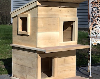 May include: A wooden cat house with a slanted roof and two entrances. The house is made of natural wood and has a light brown color.