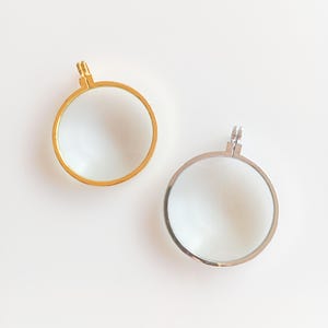 Puede incluir: Dos lupas redondas, una dorada y otra plateada, sobre un fondo blanco. Cada lupa tiene una lente circular y un pequeño lazo en la parte superior para colgar o sujetar. La dorada está a la izquierda. Ideal para manualidades o lectura.