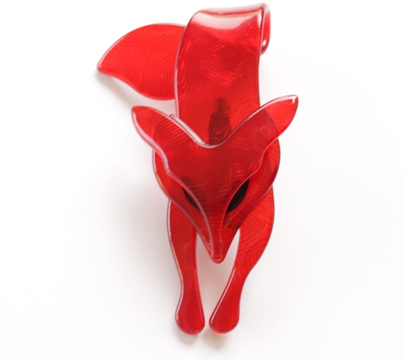 Puede incluir: Un broche de zorro de pl&aacute;stico rojo. El zorro est&aacute; en posici&oacute;n de cuclillas con la cabeza inclinada hacia abajo. El broche tiene una superficie texturizada.