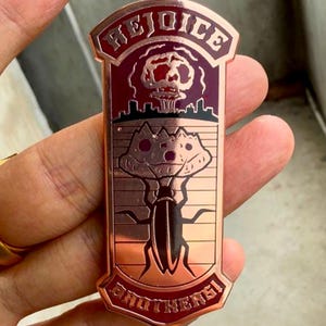 Puede incluir: Un pin rectangular de color cobre con las palabras "REJOICE BROTHERS" y una ilustración de dibujos animados de una criatura felina y una nube en forma de hongo. El pin se sostiene en una mano.