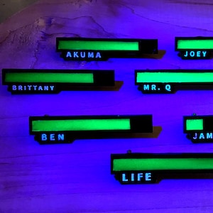 Puede incluir: Una tabla de madera con ocho etiquetas de nombre que brillan en la oscuridad. Los nombres son: Akuma, Joey, Brittany, Mr. Q, Navigator, Ben, Jamie y Life.