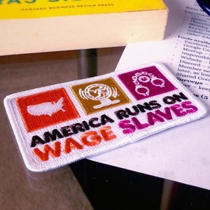 May include: A white patch with the text "America Runs on Wage Slaves" in black and pink lettering. The patch has three smaller patches on it: a red patch with a white outline of the United States, a gold patch with a black outline of a radio antenna, and a pink patch with a black outline of handcuffs.