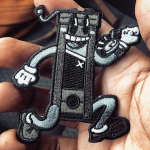 May include: A black and white embroidered patch of a cartoon character running. The character is a personified guillotine with a smiling face and a black and white striped shirt. The character is wearing black shoes and has a black and white striped hat.