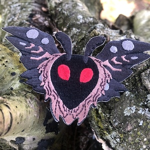 Op de afbeelding: Een geborduurde mottenpatch in zwart en roze met rode ogen. De mot heeft een pluche textuur en heeft de vorm van een hart.