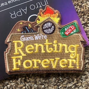 May include: A brown and yellow embroidered patch with the text "Guess We're Renting Forever!"  The patch features a house with a chimney on fire, a sign that says "Sold", and stars in the sky.