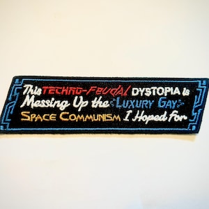 May include: A rectangular black embroidered patch with a blue circuit board border. The text reads: This TECHNO-FEUDAL DYSTOPIA is Messing Up the LUXURY GAY SPACE COMMUNISM I Hoped for, featuring white, red, blue, and yellow thread colours.