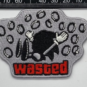 May include: A gray embroidered patch featuring a black silhouette of a character lying face down surrounded by white circular outlines. The word "WASTED" is stitched in bold red capital letters at the bottom. A black ruler shows the patch is approximately 2.5 inches wide.