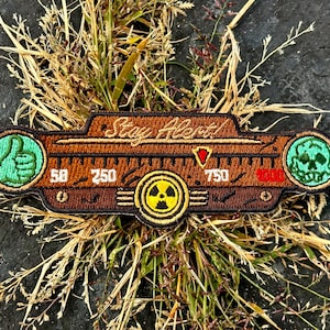 Può includere: Una toppa marrone e nera con la scritta "Stay Alert!" e un simbolo di radiazione. La toppa presenta disegni verdi di pollice in su e teschio, insieme a indicatori numerici. La toppa è circondata dall'erba.