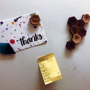 May include: A white refrigerator with a yellow sticky note that says "Shopping" and a list of groceries. There is also a card with the word "thanks" written on it, a blue diamond-shaped magnet, and a wooden magnet with the word "love" on it.