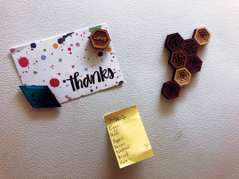 May include: A white refrigerator with a yellow sticky note that says "Shopping" and a list of groceries. There is also a card with the word "thanks" written on it, a blue diamond-shaped magnet, and a wooden magnet with the word "love" on it.