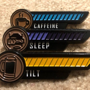 Könnte beinhalten: Drei schwarze Metallnadeln mit unterschiedlich farbigen Balken und Symbolen. Die obere Nadel hat einen blauen Balken und ein Kaffeetassen-Symbol mit dem Wort "CAFFEINE". Die mittlere Nadel hat einen lila Balken und ein Bettsymbol mit dem Wort "SLEEP". Die untere Nadel hat einen gelben Balken und ein Bierkrug-Symbol mit dem Wort "TILT".